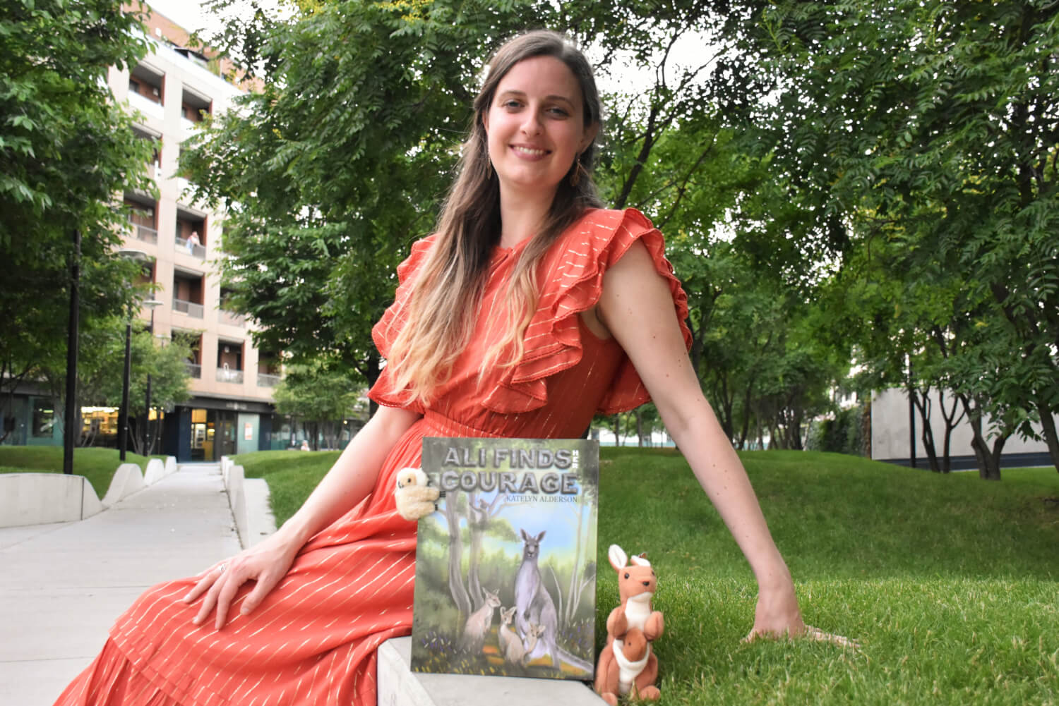 Katelyn is sitting in the park wearing an orange dress, with her book
